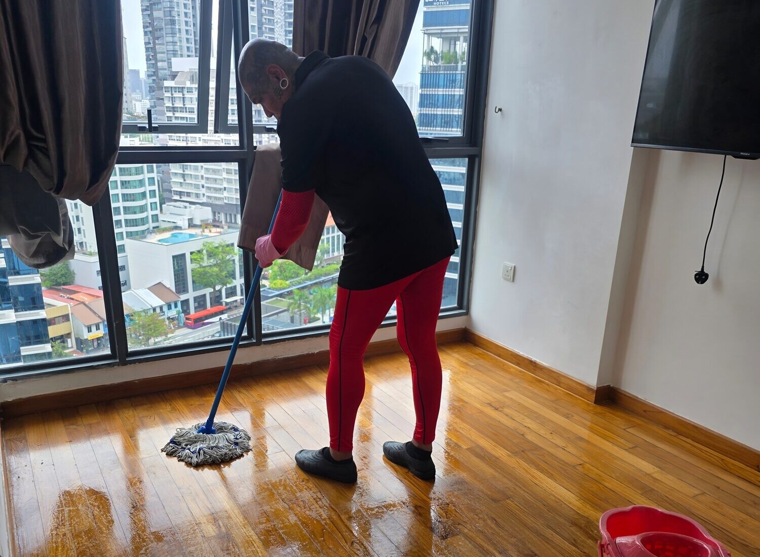 A worker in a black top and red leggings mops a wet wooden floor in an empty high-rise apartment, with a mop bucket in the foreground and a city view through the windows.